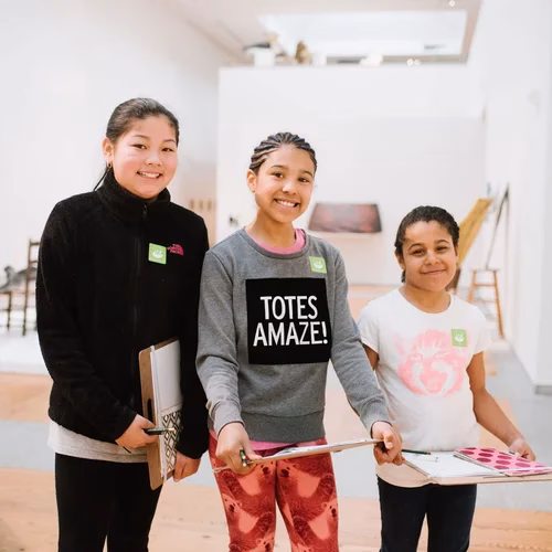 Three girls with art supplies smiling in a gallery.